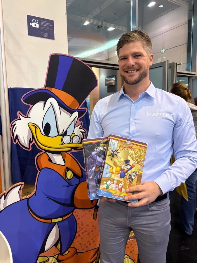 A Maestro Media team member holding the REWIND editions of Disney's Gargoyles and DuckTales beside a Scrooge McDuck standee.